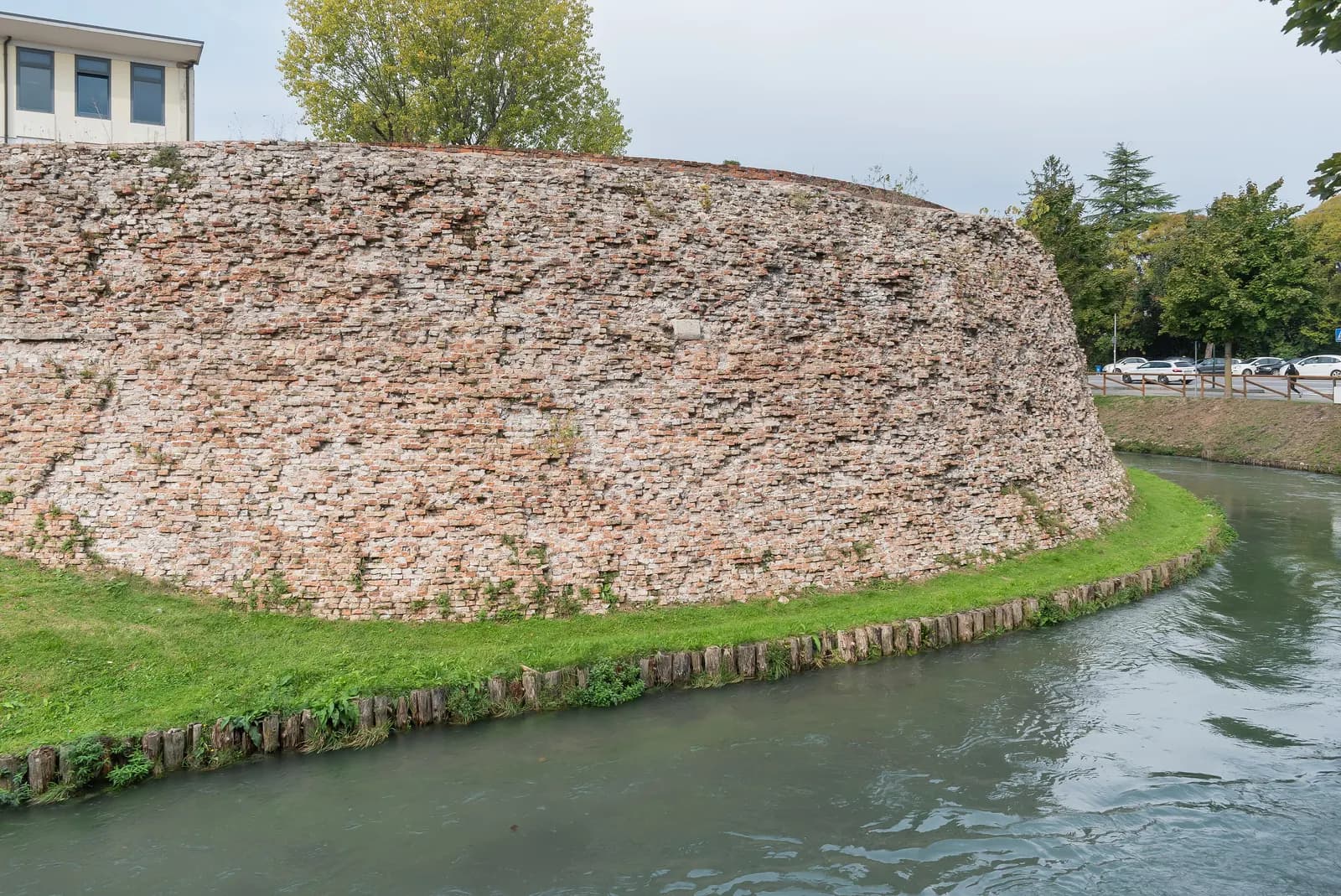 A rounded brick bastion of the sixteenth-century walls of Treviso rising from a grass bank above the moat, with a road and parked cars behind it.