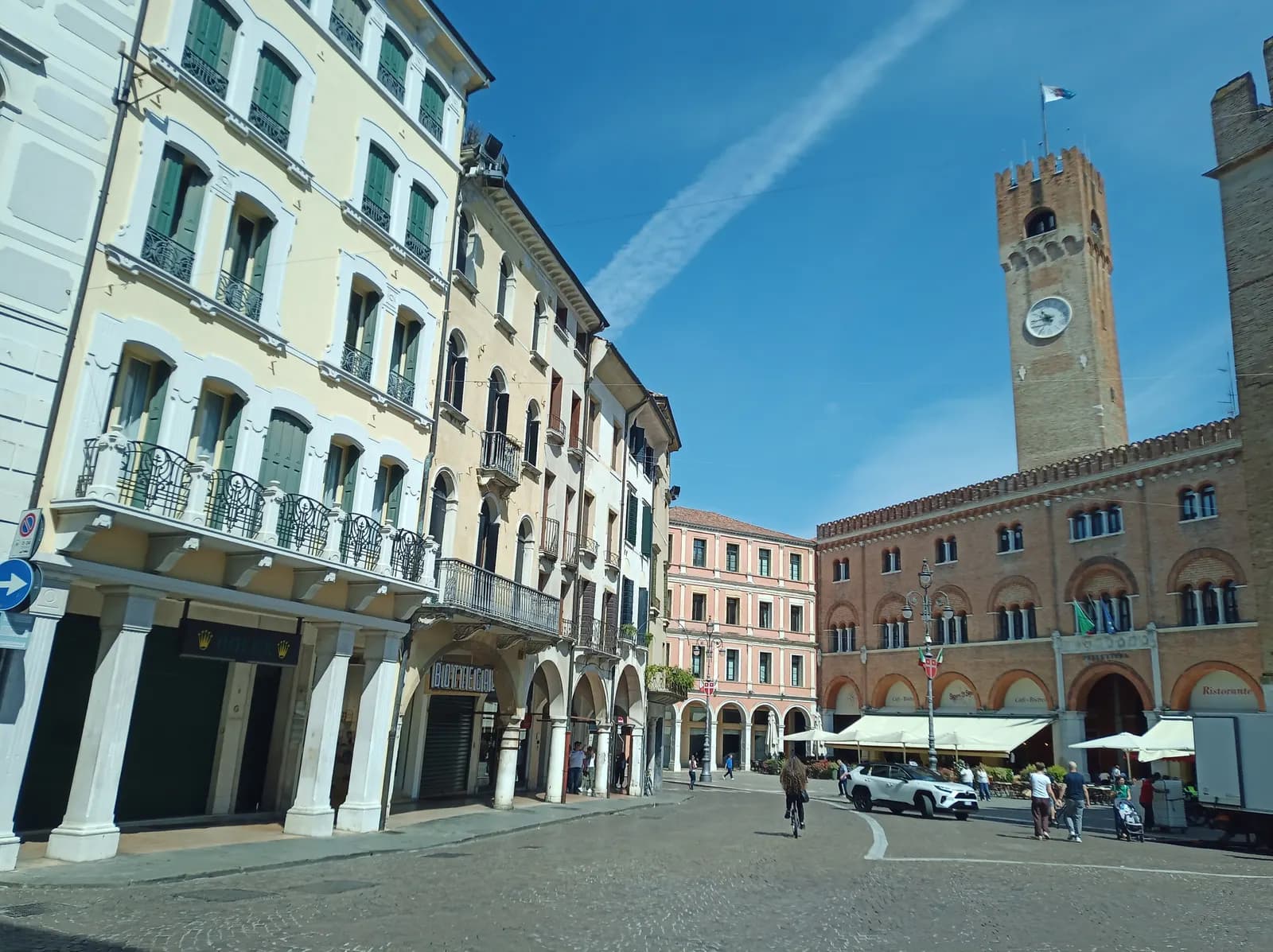 Piazza dei Signori in Treviso under a blue sky, with the brick Palazzo dei Trecento, the civic clock tower, arcaded shopfronts, cafe awnings and a car crossing the cobbles.