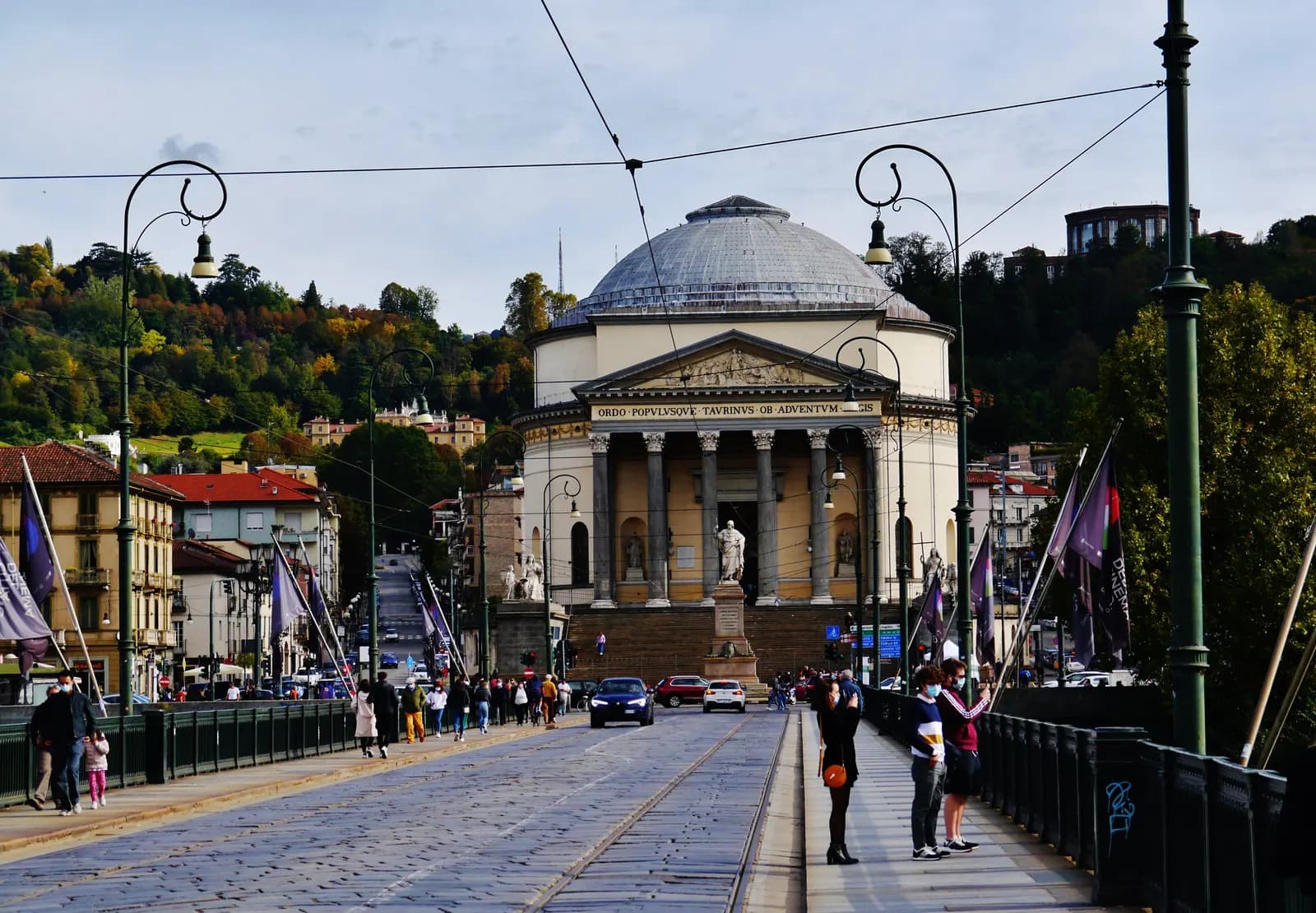 The domed, columned church of the Gran Madre di Dio in Turin seen along the bridge over the Po, with tram tracks, cars and the wooded hill behind it.