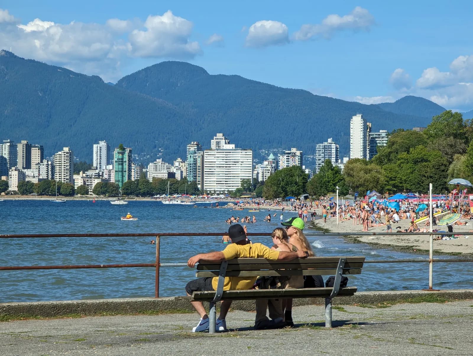 People on a bench above a busy Kitsilano Beach, looking across English Bay to the West End towers and the North Shore mountains