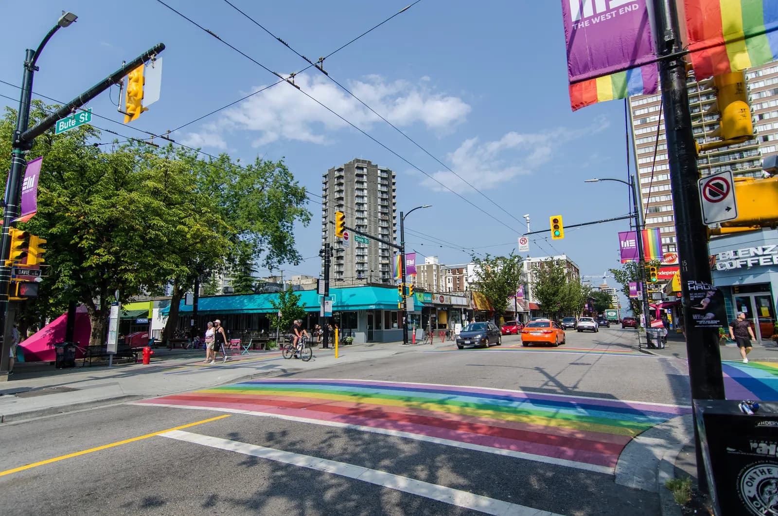 The corner of Davie Street and Bute Street in the West End, with a rainbow crosswalk, cafés, trolley bus wires and apartment towers