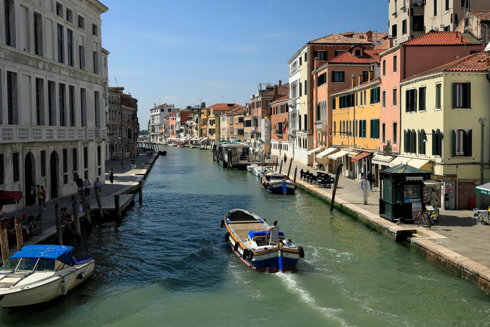 The Canale di Cannaregio seen from the Ponte delle Guglie, with a work boat, moored motorboats, the Guglie vaporetto stop and pastel houses with café tables along the fondamenta.