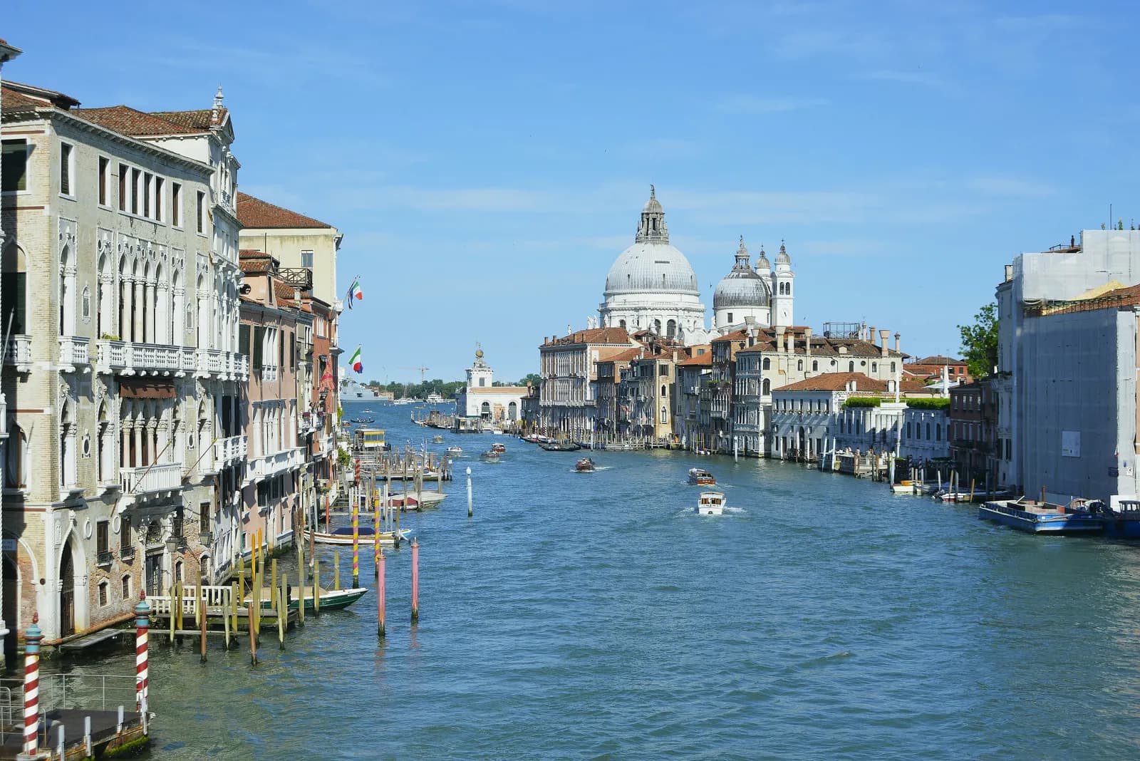 The Grand Canal seen from the Accademia bridge on a clear day, with palaces and striped mooring poles on the left bank, motorboats on the water and the domes of Santa Maria della Salute at the end.