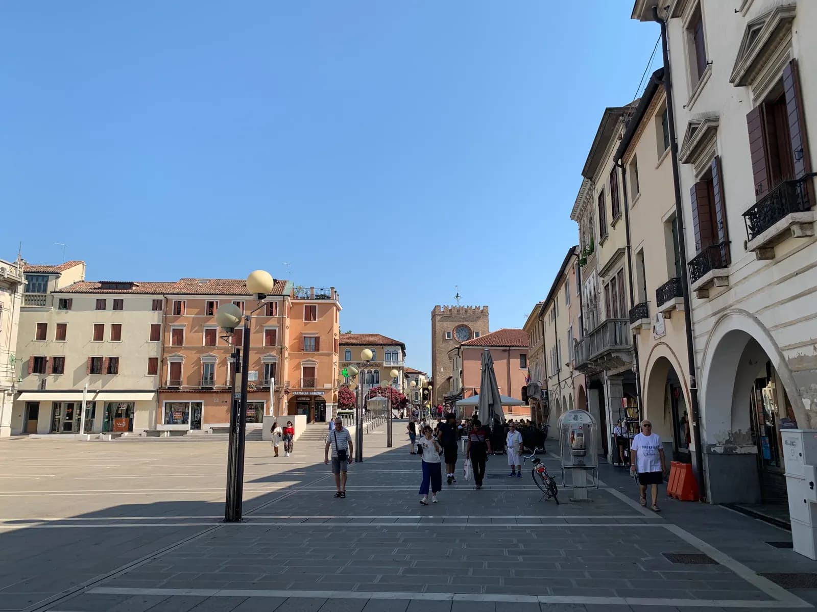 Piazza Ferretto in Mestre on a sunny morning, with arcaded buildings on the right, a café parasol and the brick Torre di Mestre clock tower at the far end.