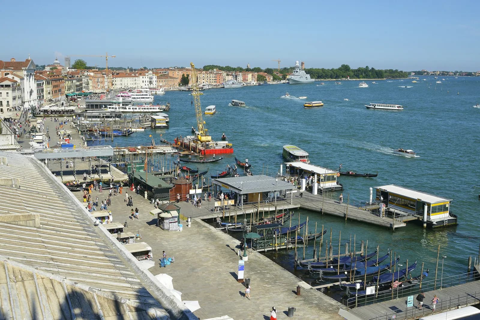 The Riva degli Schiavoni seen from above, with gondola moorings and vaporetto piers along the quay, boats on the Bacino di San Marco and the Castello shore beyond.