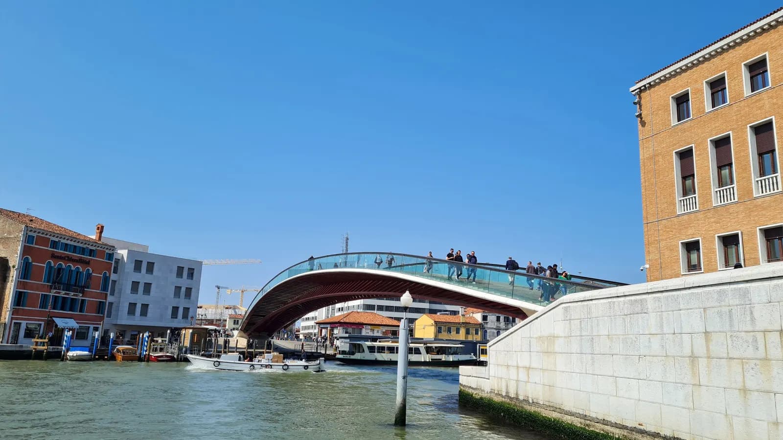 The glass and steel Ponte della Costituzione arching over the Grand Canal between the railway station and Piazzale Roma, with people crossing and boats moored below.