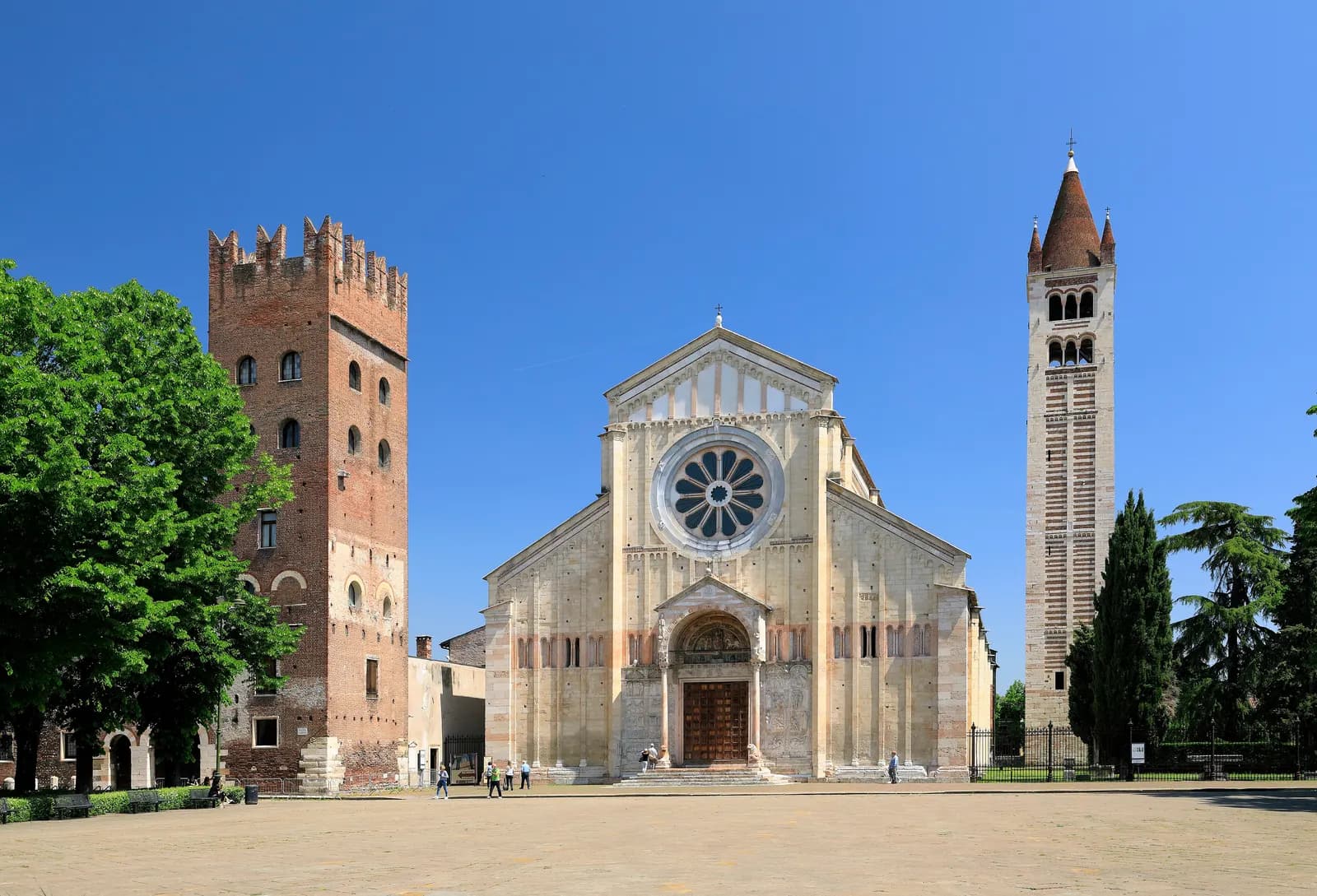 The Romanesque Basilica di San Zeno with its rose window, a crenellated brick tower on the left and the striped bell tower on the right, across the open Piazza San Zeno.