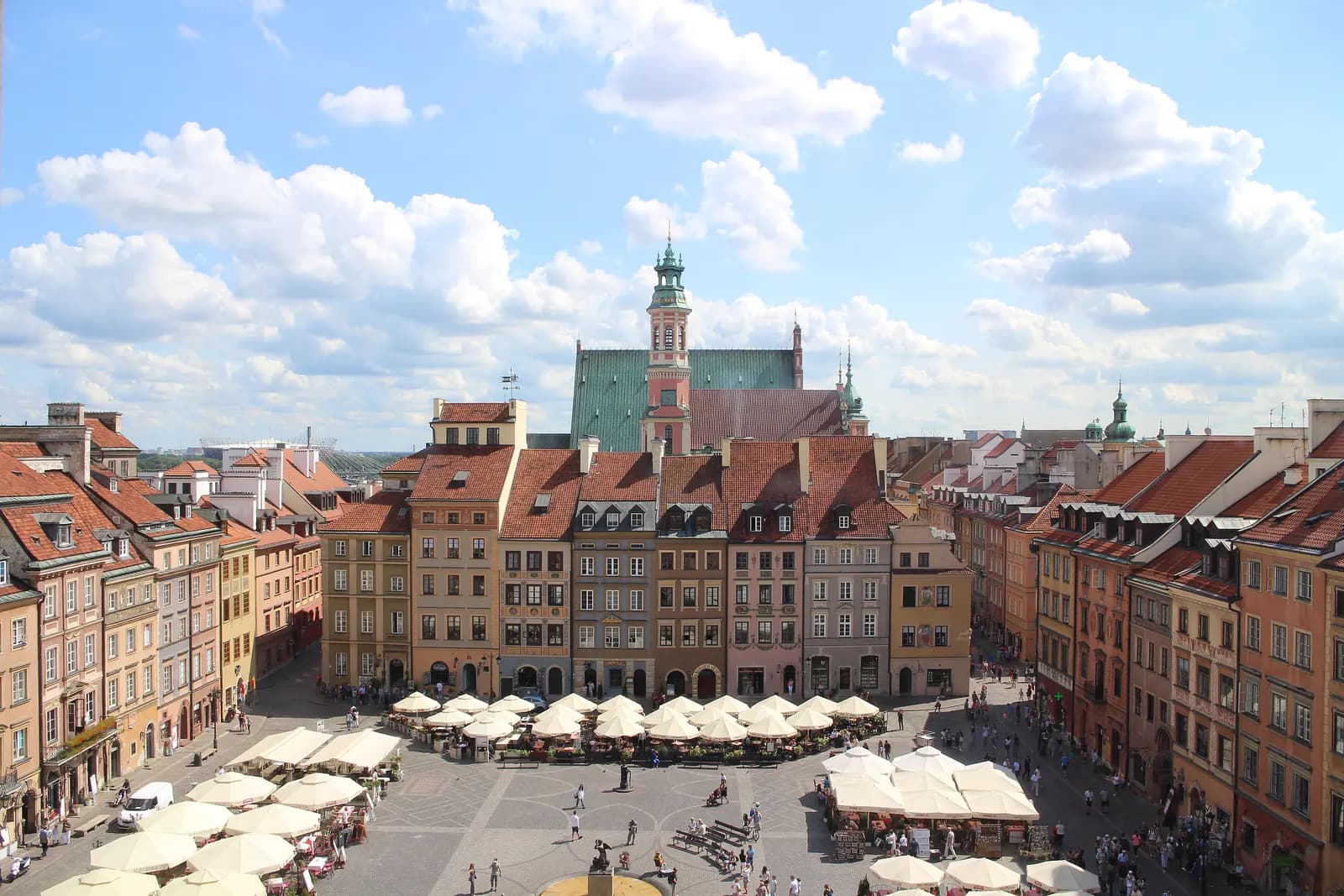Warsaw's Old Town Market Square seen from above, with rows of tall coloured townhouses, café umbrellas on the square and a church tower behind