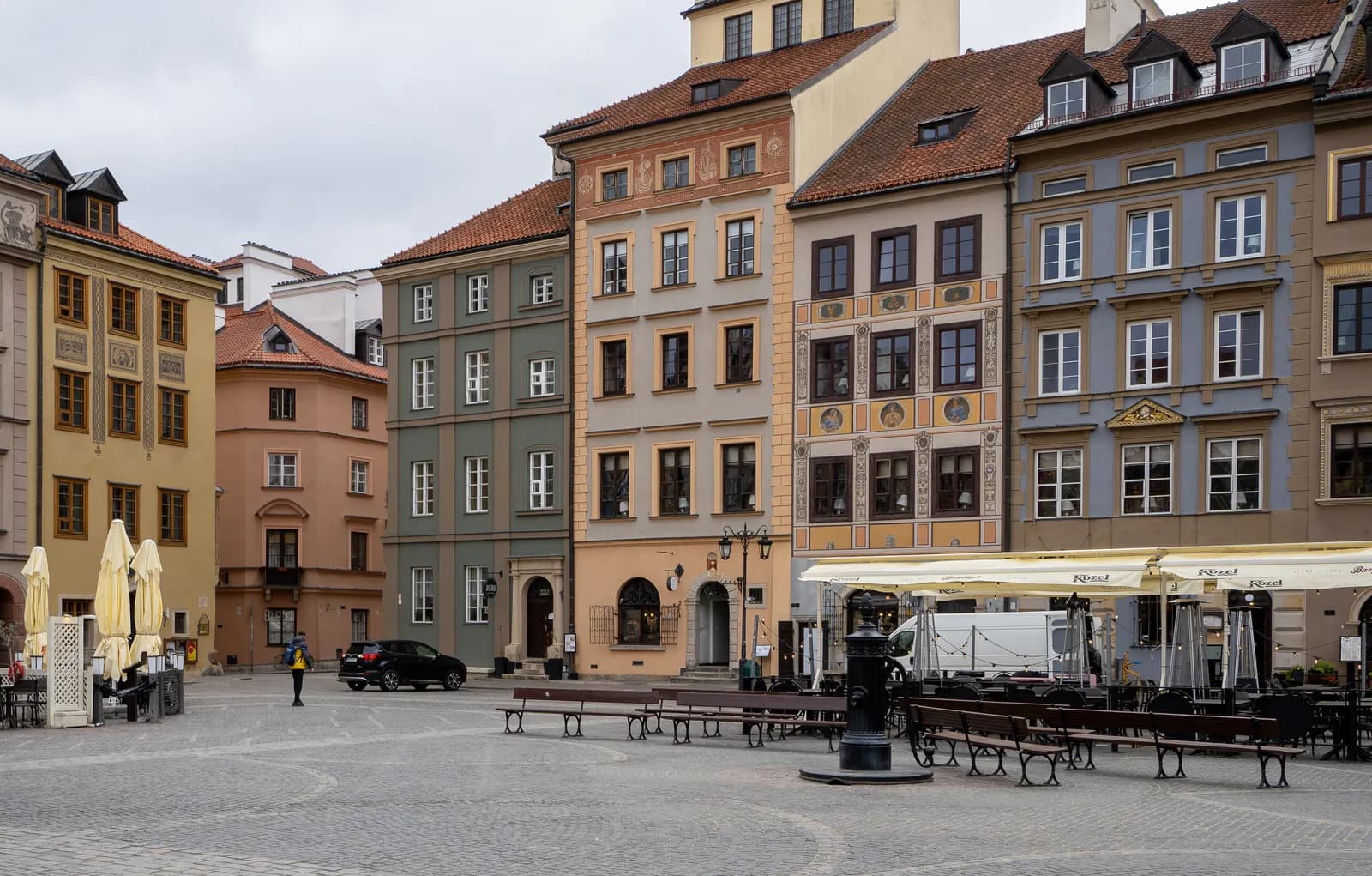 Coloured townhouses with steep tiled roofs on one side of Warsaw's Old Town Market Square, with benches and café awnings on the cobbles