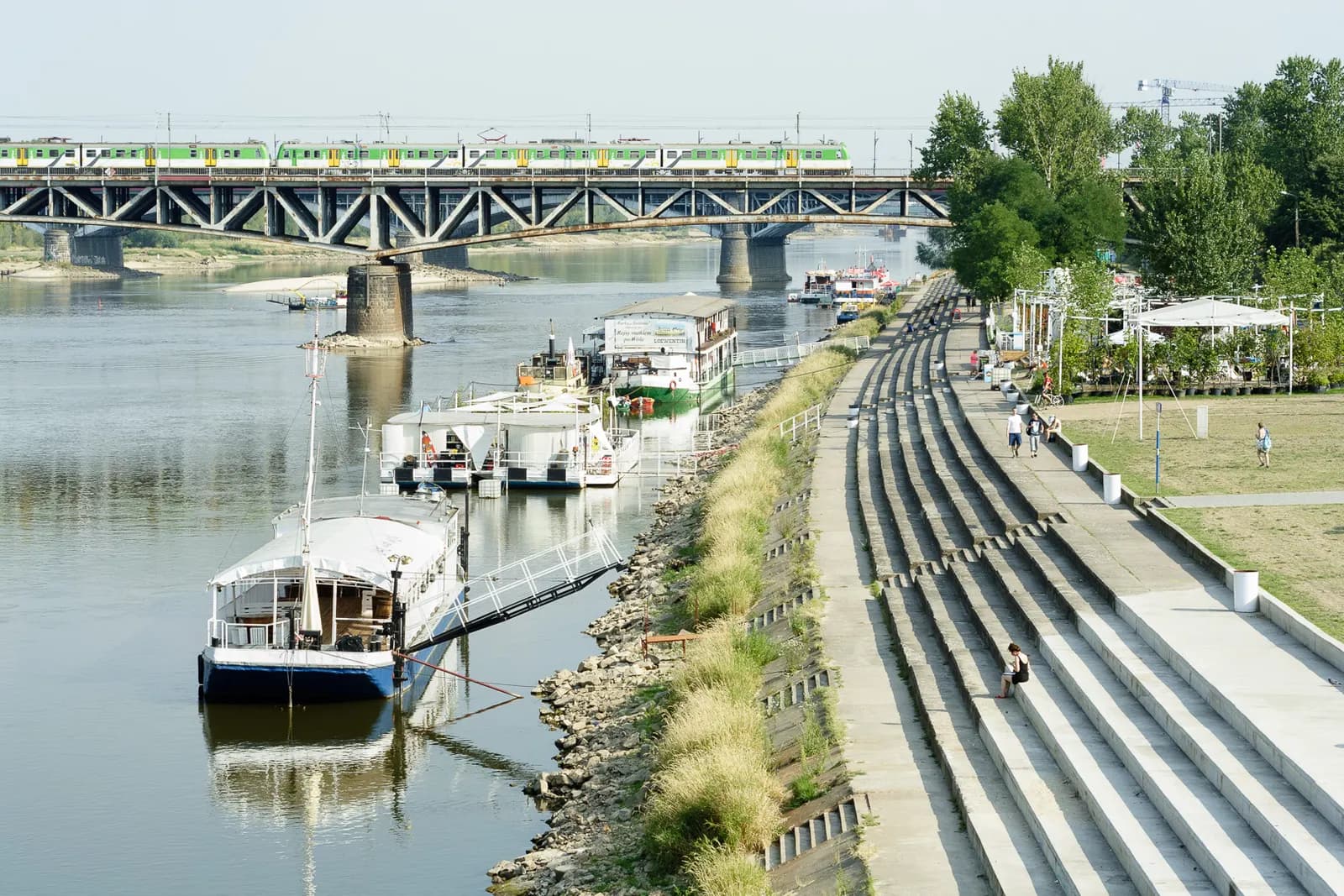 The Vistula boulevards at Powiśle in Warsaw, with stepped embankment seating, moored boats and a railway bridge across the river