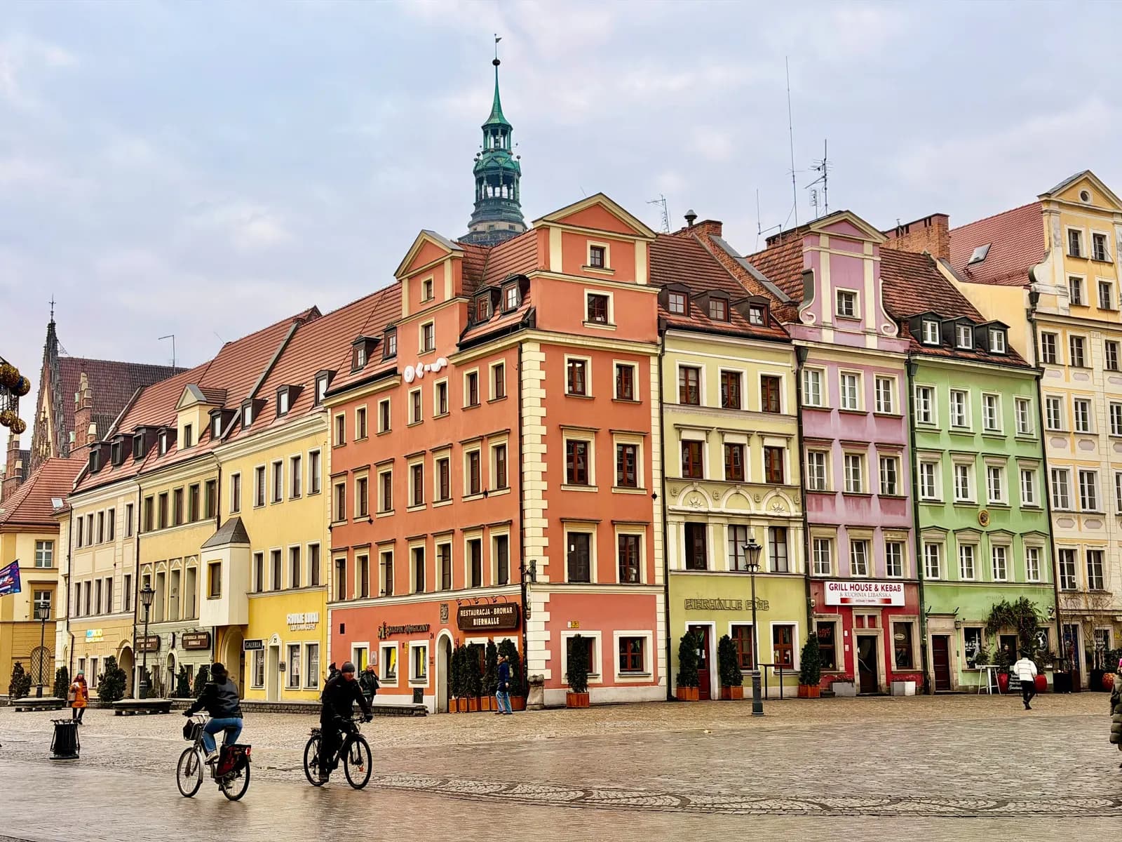 A row of colourful gabled houses in the Market Square in Wrocław, with the Town Hall tower rising behind and two cyclists on the wet paving