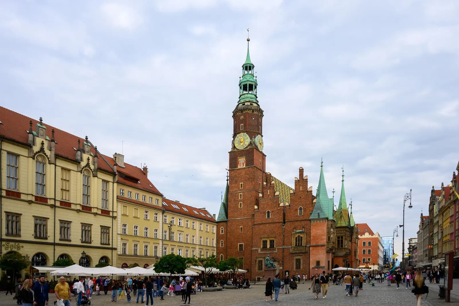 The Gothic brick Town Hall of Wrocław with its clock tower and green spires on the Market Square, with café umbrellas and people walking
