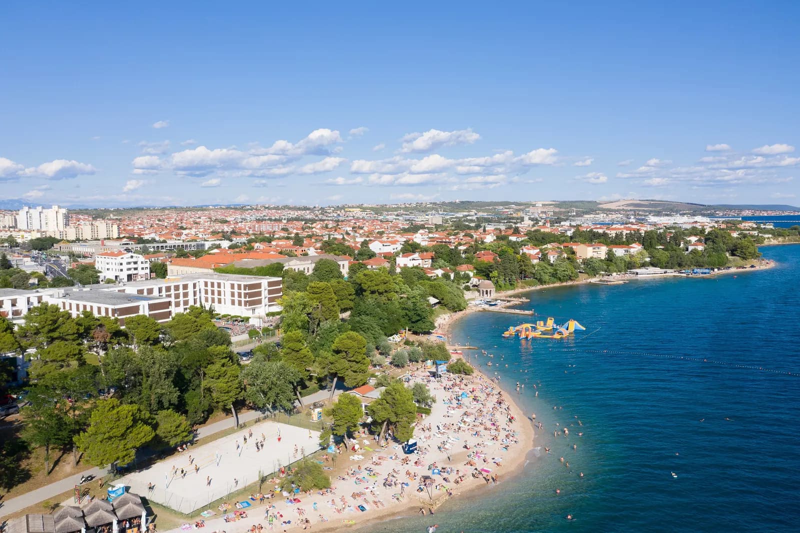 Kolovare beach in Zadar from above, with swimmers, a beach volleyball court, a hotel among pine trees and residential streets behind