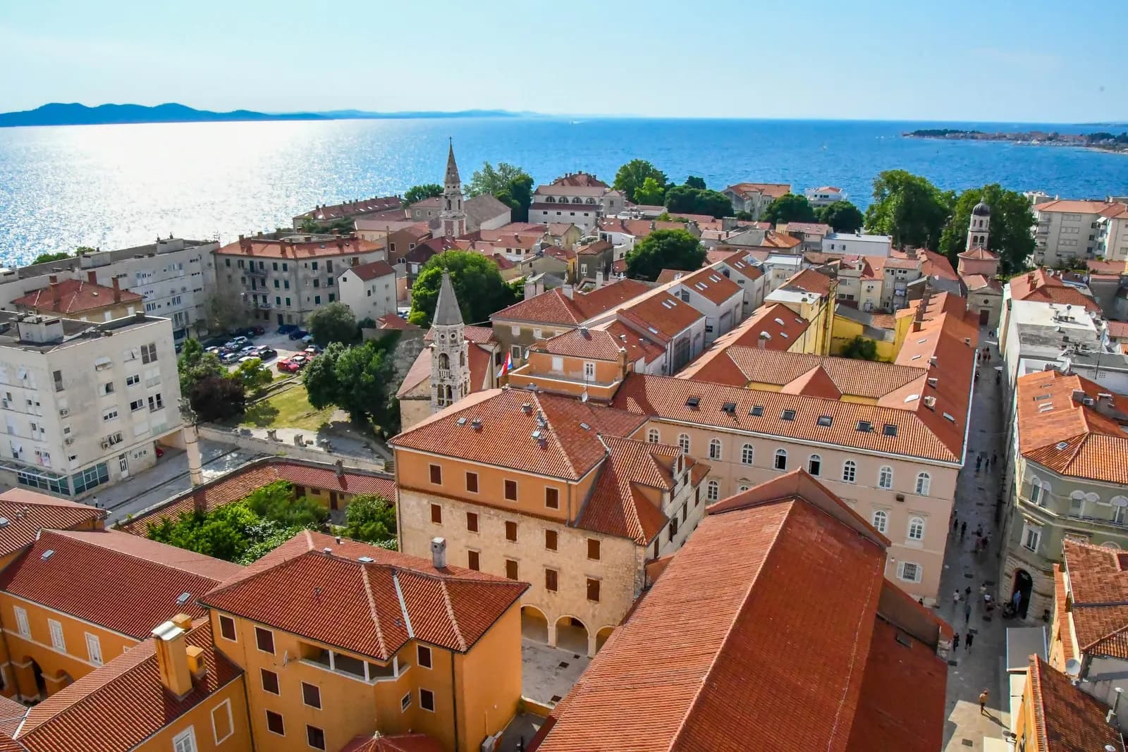 Tiled roofs, bell towers and narrow streets of Zadar Old Town seen from above, with a car park between the buildings and the sea behind