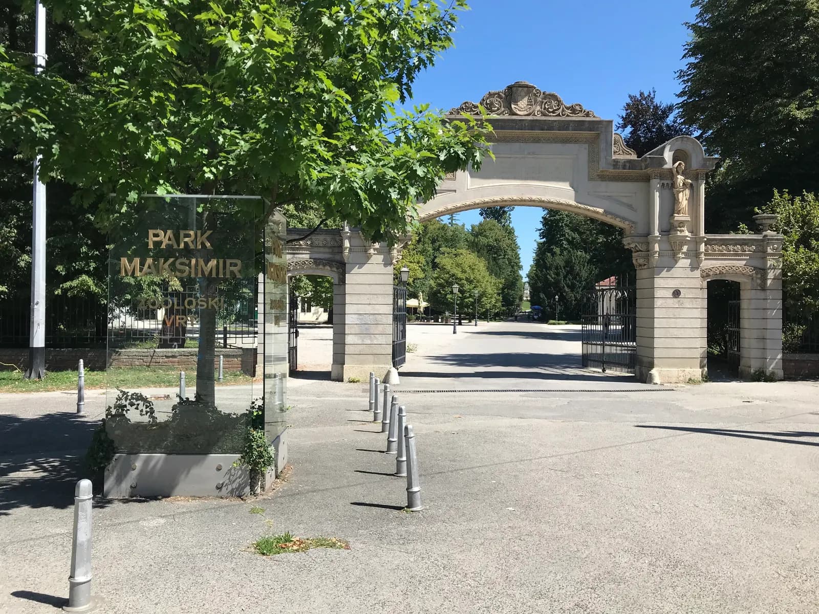 The stone entrance gate of Maksimir park in Zagreb, with a glass sign for the park and the zoo and a tree-lined avenue beyond