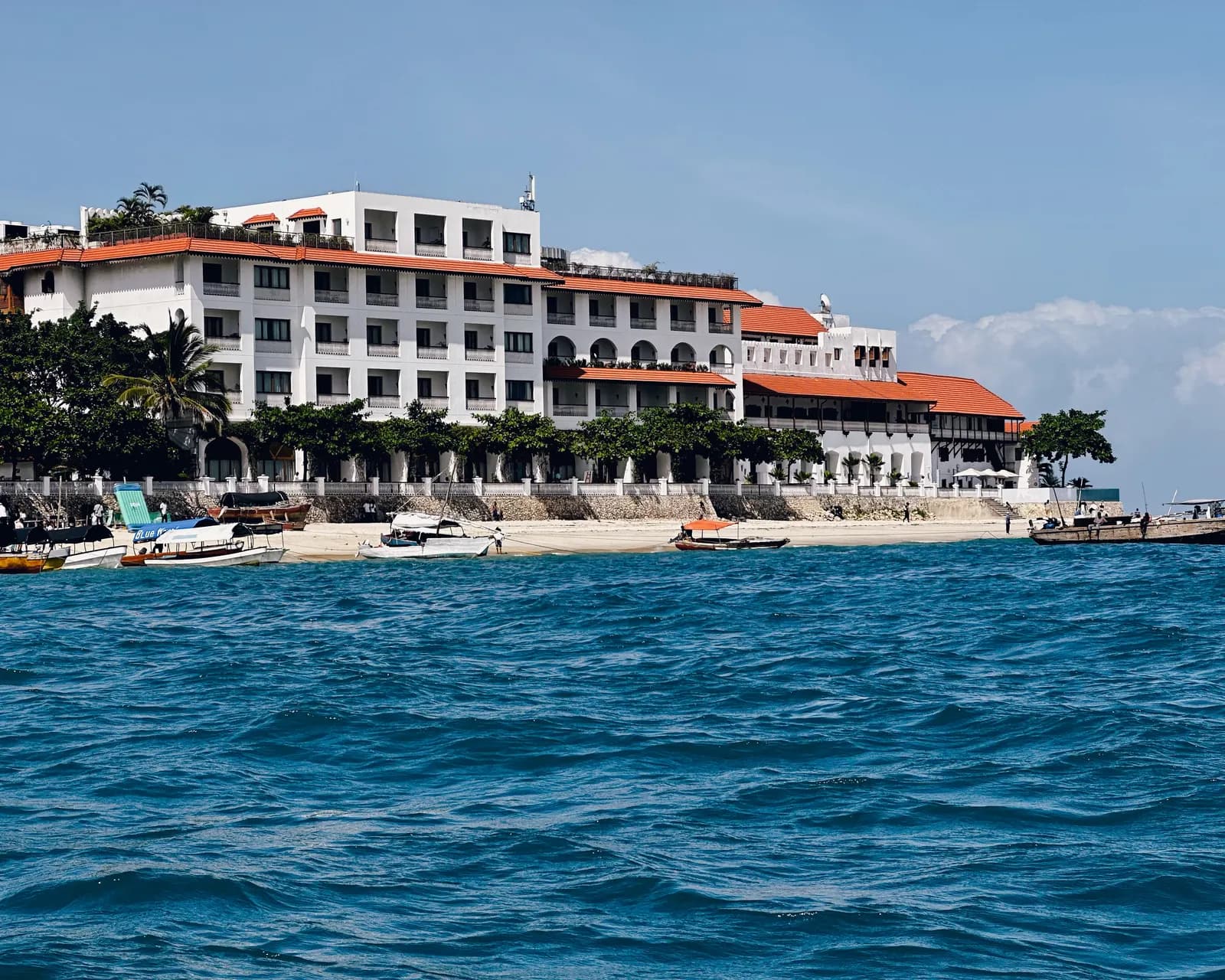 The Stone Town seafront seen from the water to the north, with a large white building with red roofs, palm trees and small boats pulled up on the beach