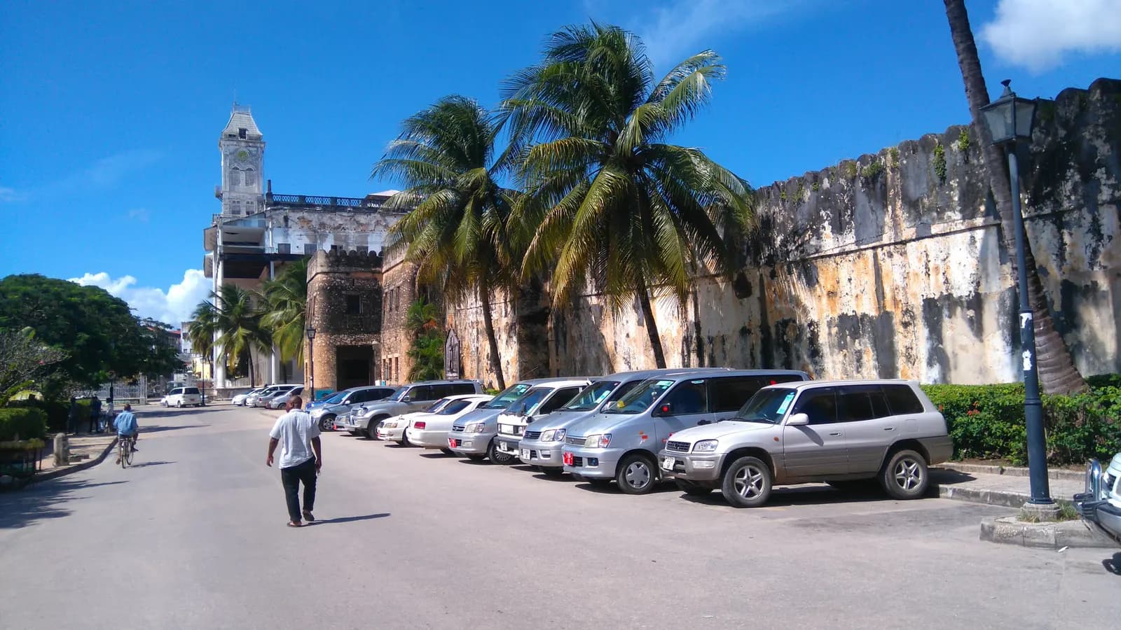 A row of cars parked along the street beside the walls of the Old Fort in Stone Town, under palm trees, with the House of Wonders tower behind